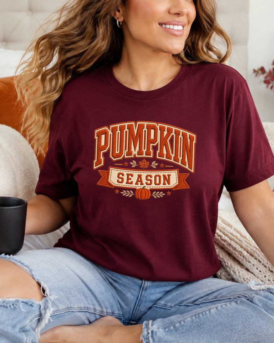 A woman wearing a maroon short-sleeve graphic T-shirt with 'PUMPKIN SEASON' printed in orange and white, paired with light-wash ripped jeans, sitting on a couch.