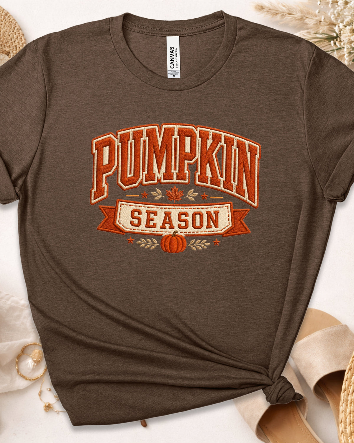 A dark brown short-sleeve t-shirt with a 'Pumpkin Season' graphic in orange and cream, styled in a flat-lay with a knot at the bottom hem, surrounded by neutral props.