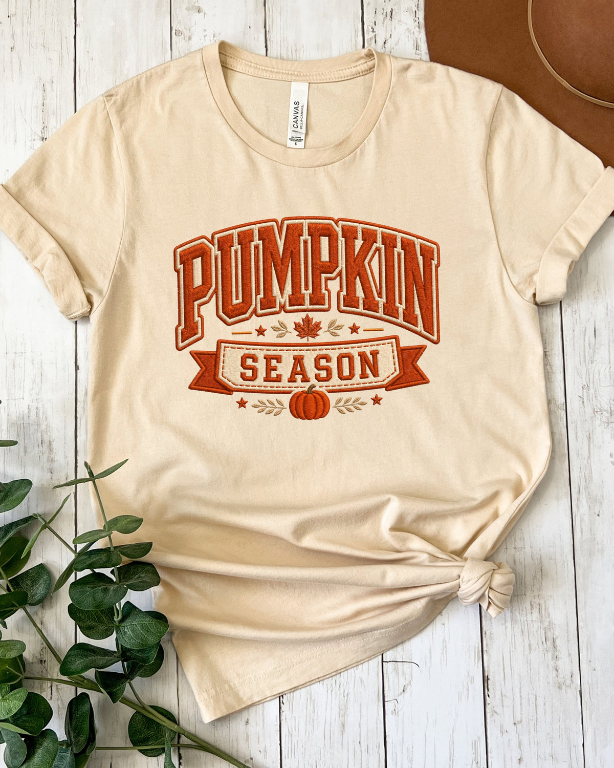 A beige short-sleeve t-shirt with a 'Pumpkin Season' graphic in orange and brown, styled on a white wood plank background with a knot at the bottom hem.