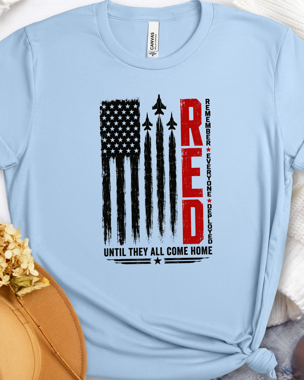 A light blue short-sleeve t-shirt with a patriotic graphic on the front. The graphic features a distressed black American flag with fighter jets flying through it, the word 'RED' in bold red letters, and the text 'UNTIL THEY ALL COME HOME' in black.