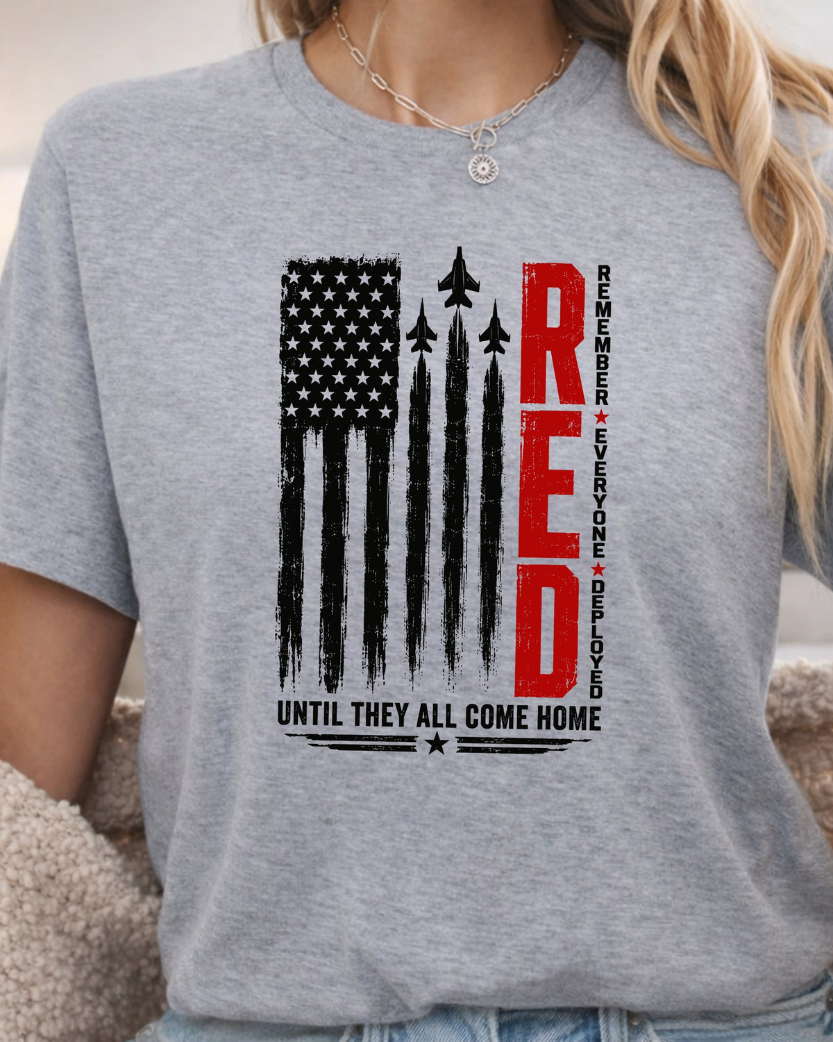 A woman wearing a heather gray short-sleeve t-shirt with a patriotic graphic on the front. The graphic features a distressed black American flag, fighter jets, the word 'RED' in bold red letters, and the text 'UNTIL THEY ALL COME HOME' in black.