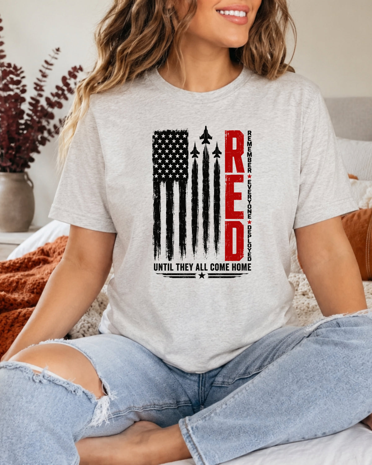 A woman wearing a light heather gray graphic t-shirt with a distressed American flag design and the word 'RED' in vertical red letters, paired with ripped light blue jeans.