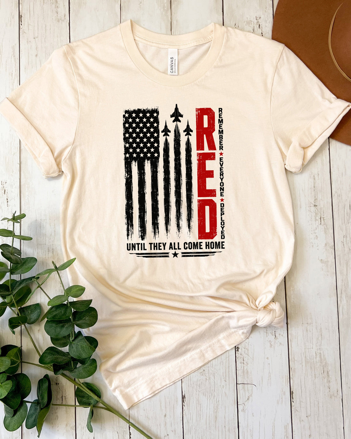 A cream-colored short-sleeve t-shirt with a patriotic graphic on the front, laid flat on a white wood plank background with green leaves and a brown hat as props.