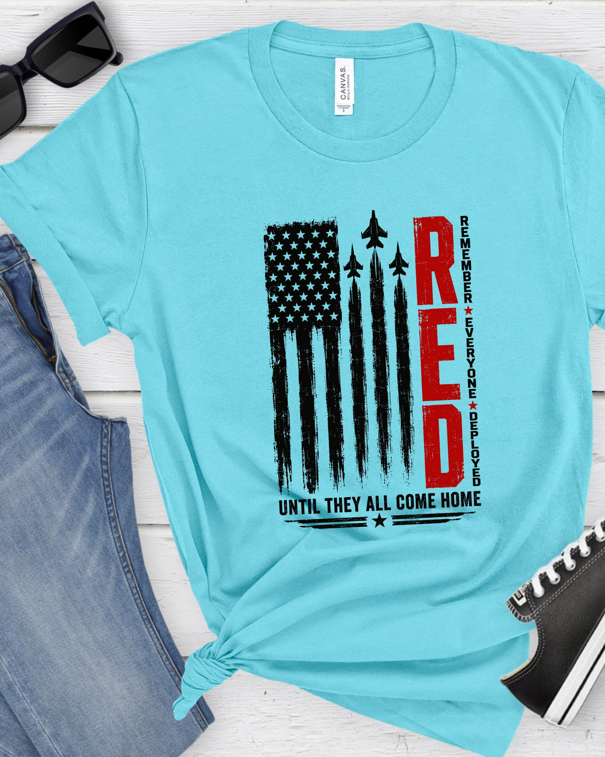 A light blue t-shirt with a patriotic graphic print featuring a distressed American flag, fighter jets, and the word 'RED' in bold red letters. The shirt is styled in a flat-lay arrangement with blue jeans, black sunglasses, and a black sneaker on a white wood background.