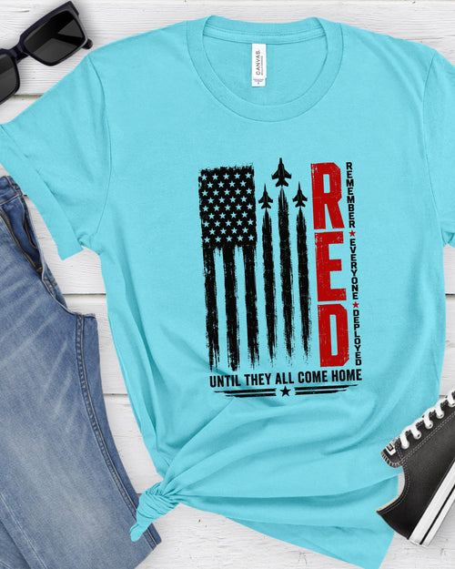 A light blue t-shirt with a patriotic graphic print featuring a distressed American flag, fighter jets, and the word 'RED' in bold red letters. The shirt is styled in a flat-lay arrangement with blue jeans, black sunglasses, and a black sneaker on a white wood background.