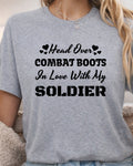 A woman wearing a heather gray t-shirt with black printed text that reads 'Head Over COMBAT BOOTS In Love With My SOLDIER' accompanied by small heart icons.