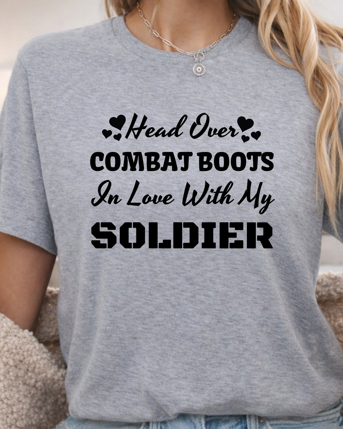 A woman wearing a heather gray t-shirt with black printed text that reads 'Head Over COMBAT BOOTS In Love With My SOLDIER' accompanied by small heart icons.