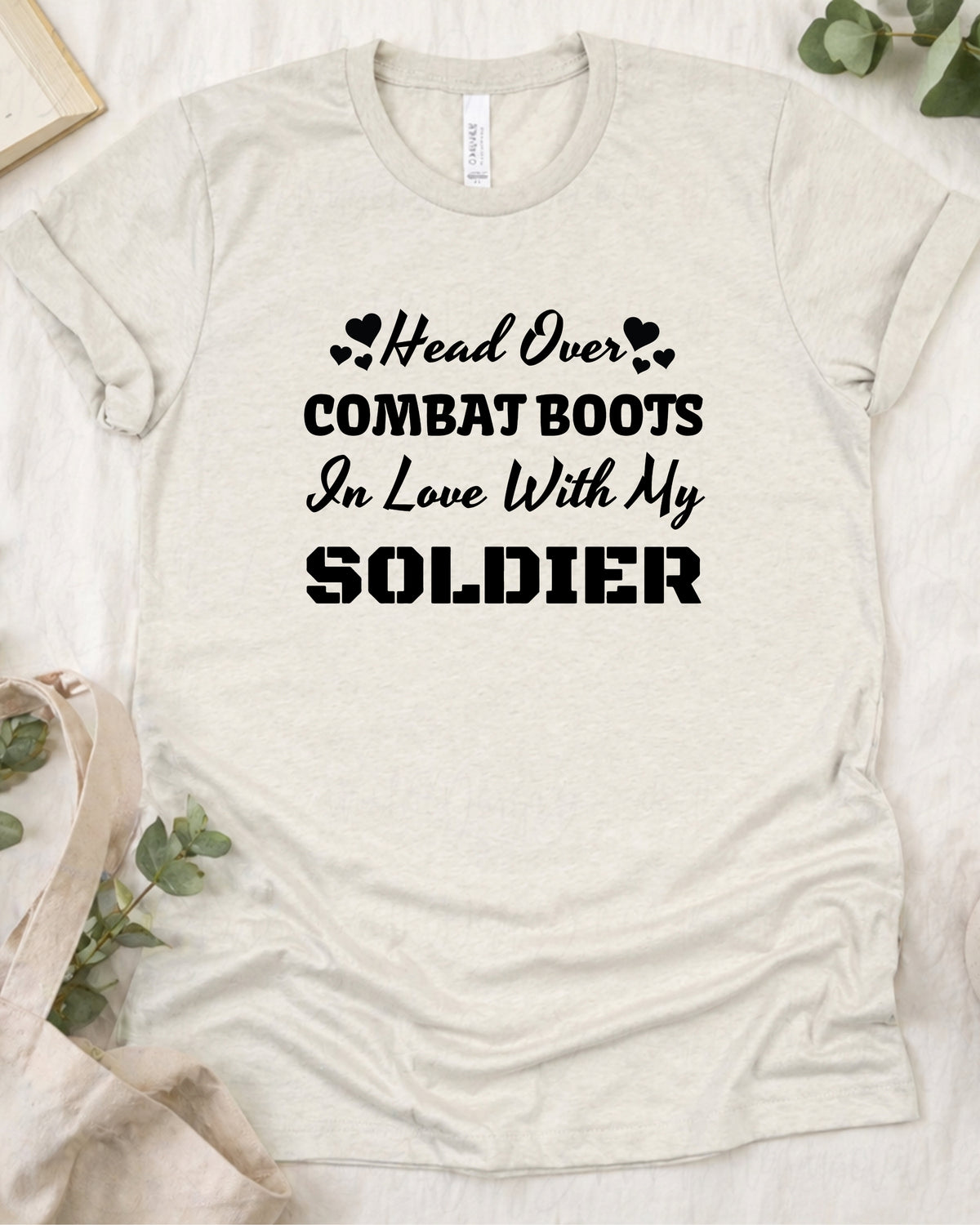 A light beige heather crewneck t-shirt with black printed text reading 'Head Over COMBAT BOOTS In Love With My SOLDIER' laid flat on a white textured background with decorative eucalyptus leaves.