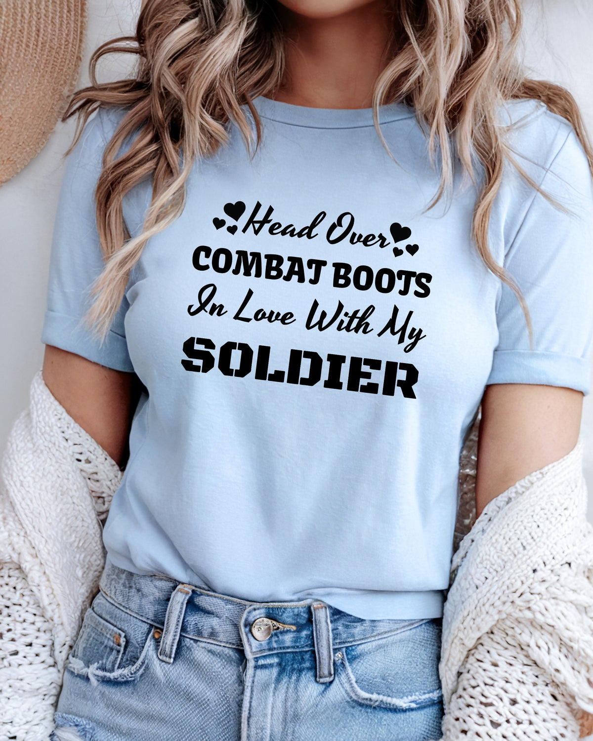 A woman wearing a light blue graphic t-shirt with the text 'Head Over COMBAT BOOTS In Love With My SOLDIER' in black lettering, styled with a white knit cardigan and denim shorts.