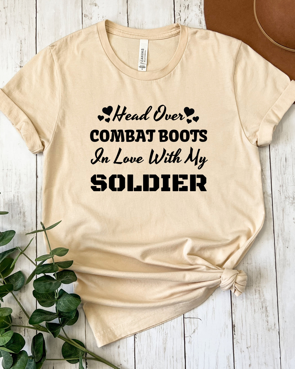 A beige graphic t-shirt with black text reading 'Head Over COMBAT BOOTS In Love With My SOLDIER' styled on a white wood plank background with a green plant and a brown hat visible.