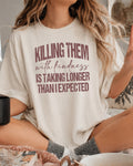 A woman wearing a cream-colored oversized graphic t-shirt with the text 'KILLING THEM with kindness IS TAKING LONGER THAN I EXPECTED' in a dusty rose-brown color. She is sitting cross-legged in a cozy indoor setting.