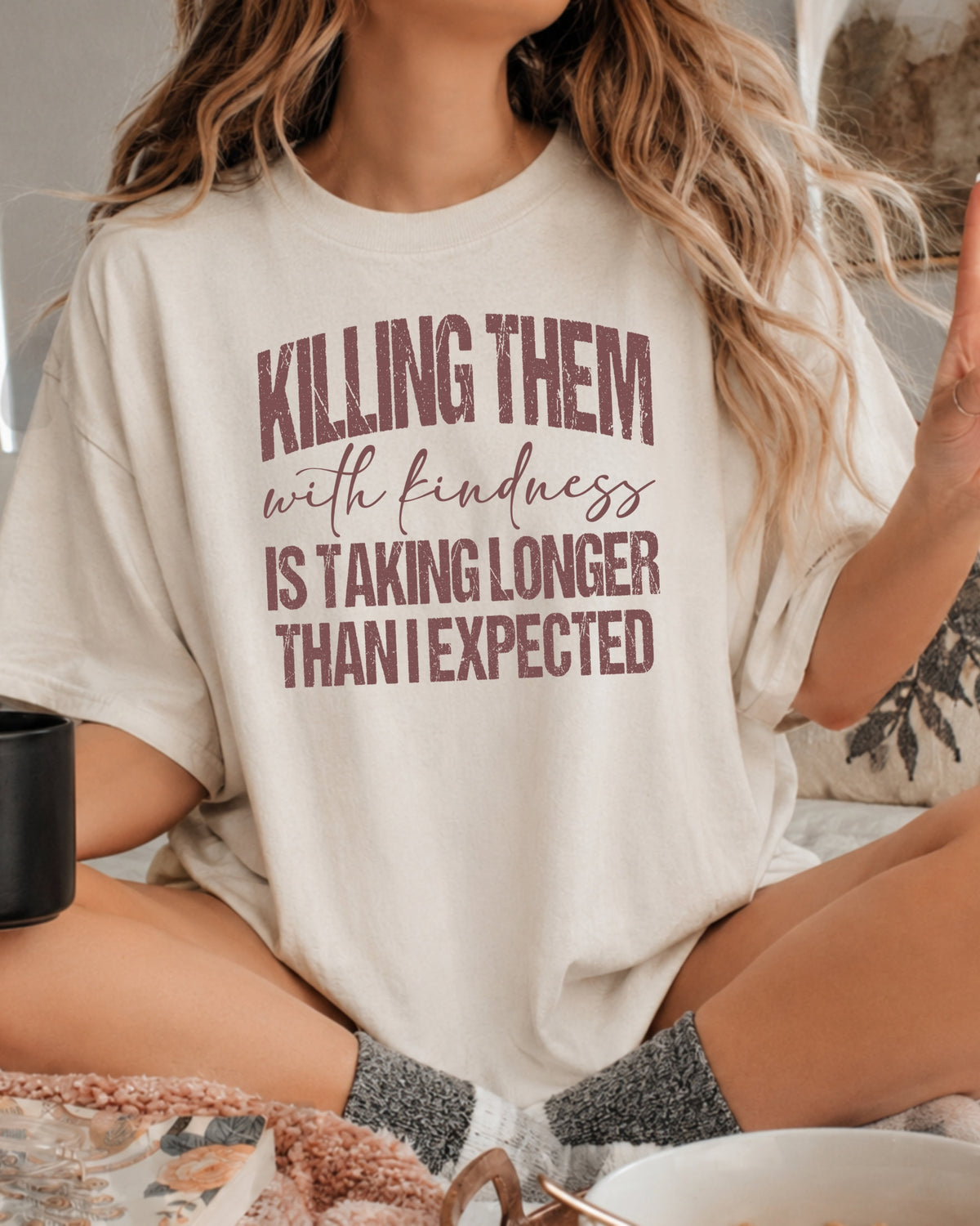 A woman wearing a cream-colored oversized graphic t-shirt with the text 'KILLING THEM with kindness IS TAKING LONGER THAN I EXPECTED' in a dusty rose-brown color. She is sitting cross-legged in a cozy indoor setting.