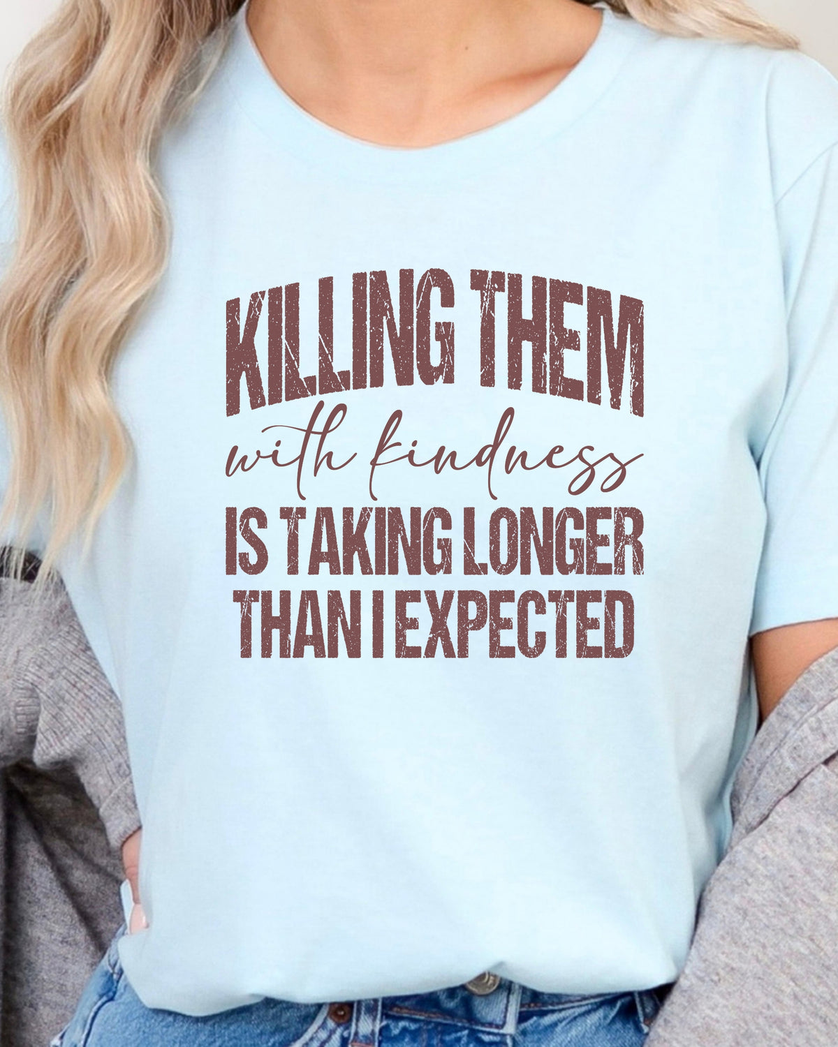 A woman wearing a light blue graphic t-shirt with the text 'KILLING THEM with kindness IS TAKING LONGER THAN I EXPECTED' in brown and black lettering, styled with a gray cardigan and blue jeans.