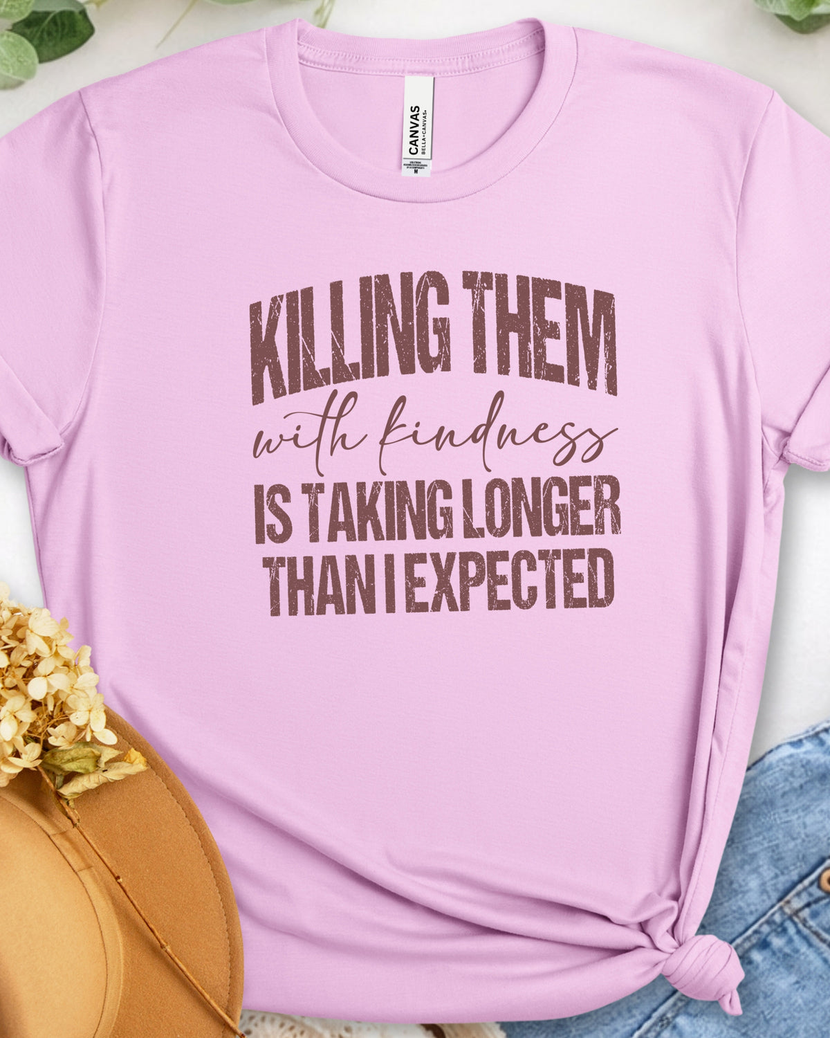 A light pink short-sleeve graphic T-shirt laid flat with a knot at the bottom right, featuring the phrase 'KILLING THEM with kindness IS TAKING LONGER THAN I EXPECTED' in brown lettering. The shirt is styled with a tan hat, blue jeans, and dried flowers on a light surface.