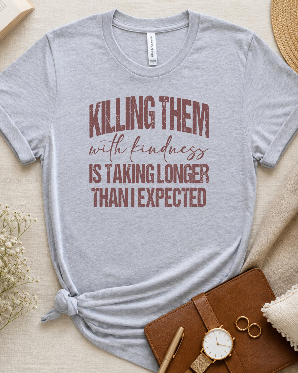 A light heather gray short-sleeve t-shirt with a distressed brown text graphic reading 'KILLING THEM with kindness IS TAKING LONGER THAN I EXPECTED'. The t-shirt is styled in a flat-lay arrangement on a beige fabric background with a brown leather notebook, a watch, rings, and dried flowers.