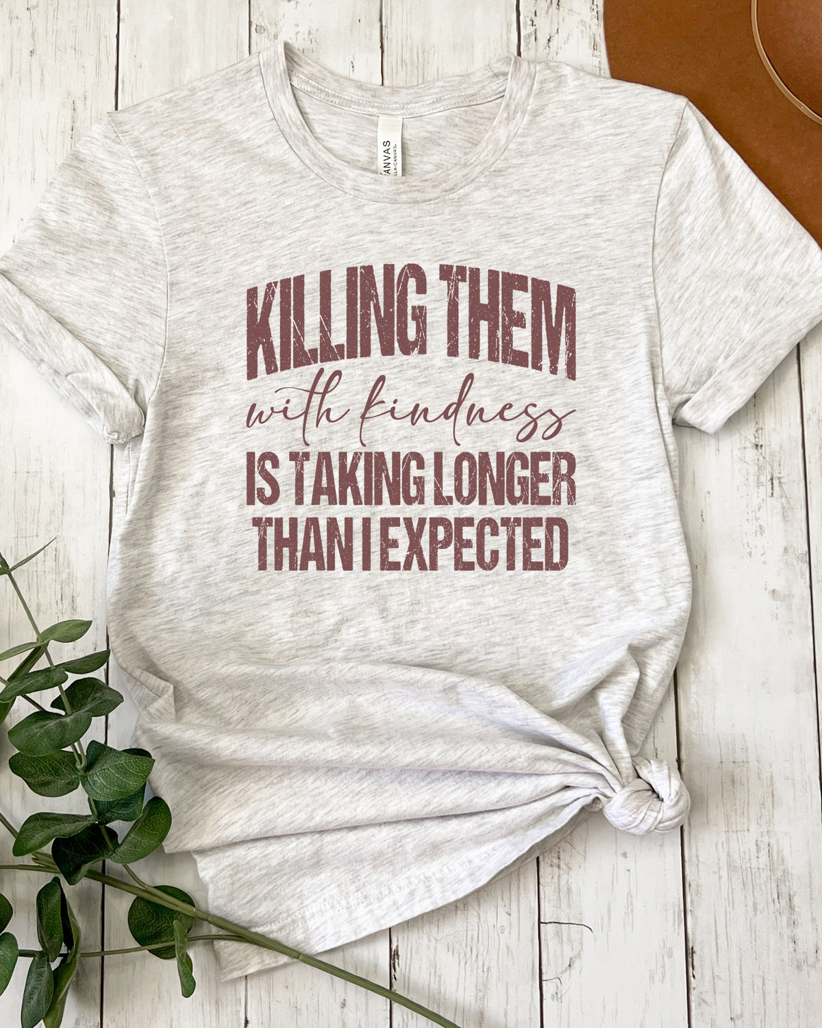 A light heather gray t-shirt with a maroon text graphic reading 'KILLING THEM with kindness IS TAKING LONGER THAN I EXPECTED', styled flat on a white wood background with a green plant and a brown hat visible.