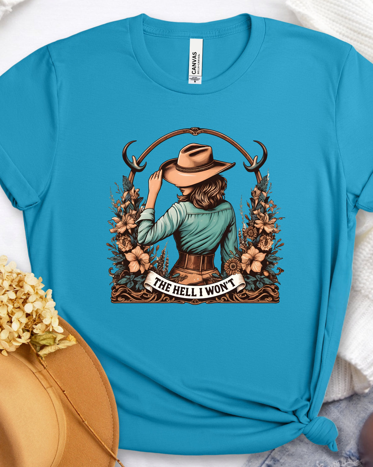 A bright turquoise blue short-sleeve t-shirt with a graphic of a woman in a cowboy hat facing away from the viewer, framed by flowers and the text 'THE HELL I WON'T'. The shirt is styled flat with a tan hat and dried flowers in the foreground.