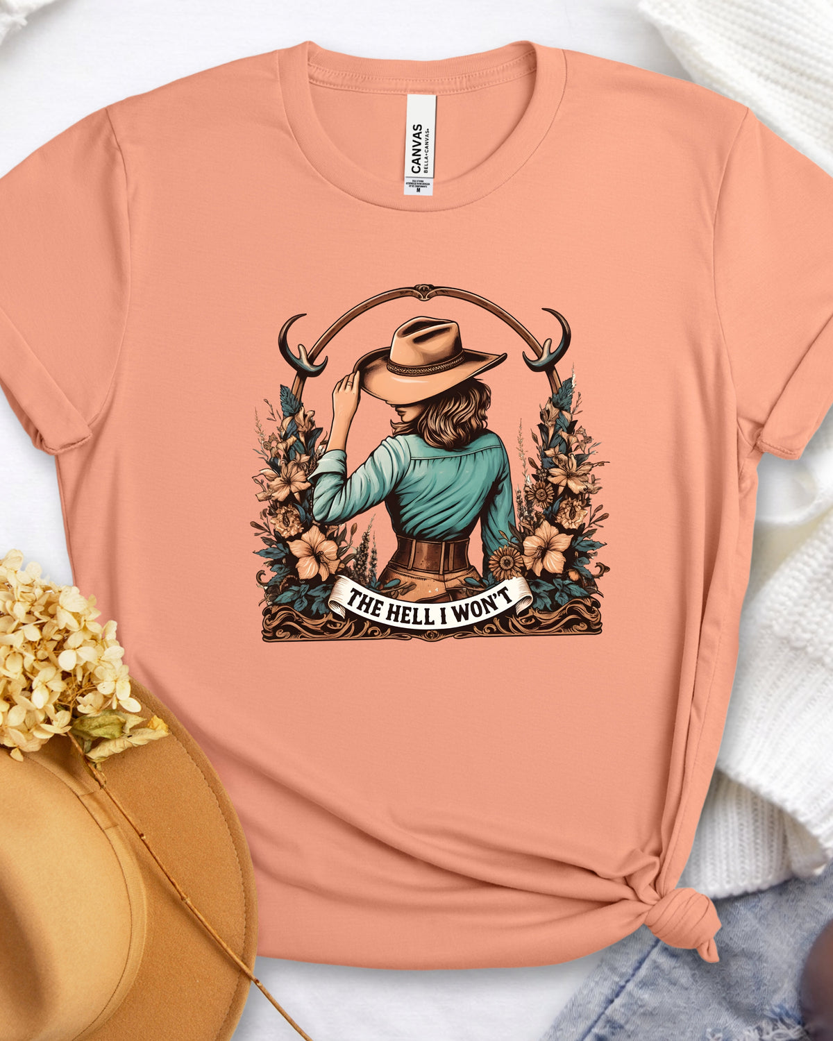 A peach-colored graphic t-shirt featuring a western-style illustration of a woman in a hat surrounded by flowers and the text 'THE HELL I WON'T'. The shirt is styled in a flat-lay arrangement with a straw hat, dried flowers, and a white knit sweater.