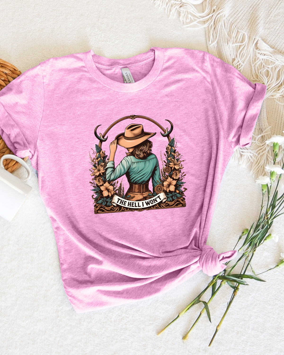 A pink short-sleeve graphic T-shirt laid flat on a light fabric surface with decorative props. The shirt has a large front print showing a seated woman in a cowboy hat surrounded by flowers and antlers, with the text 'THE HELL I WOULDN'T'.