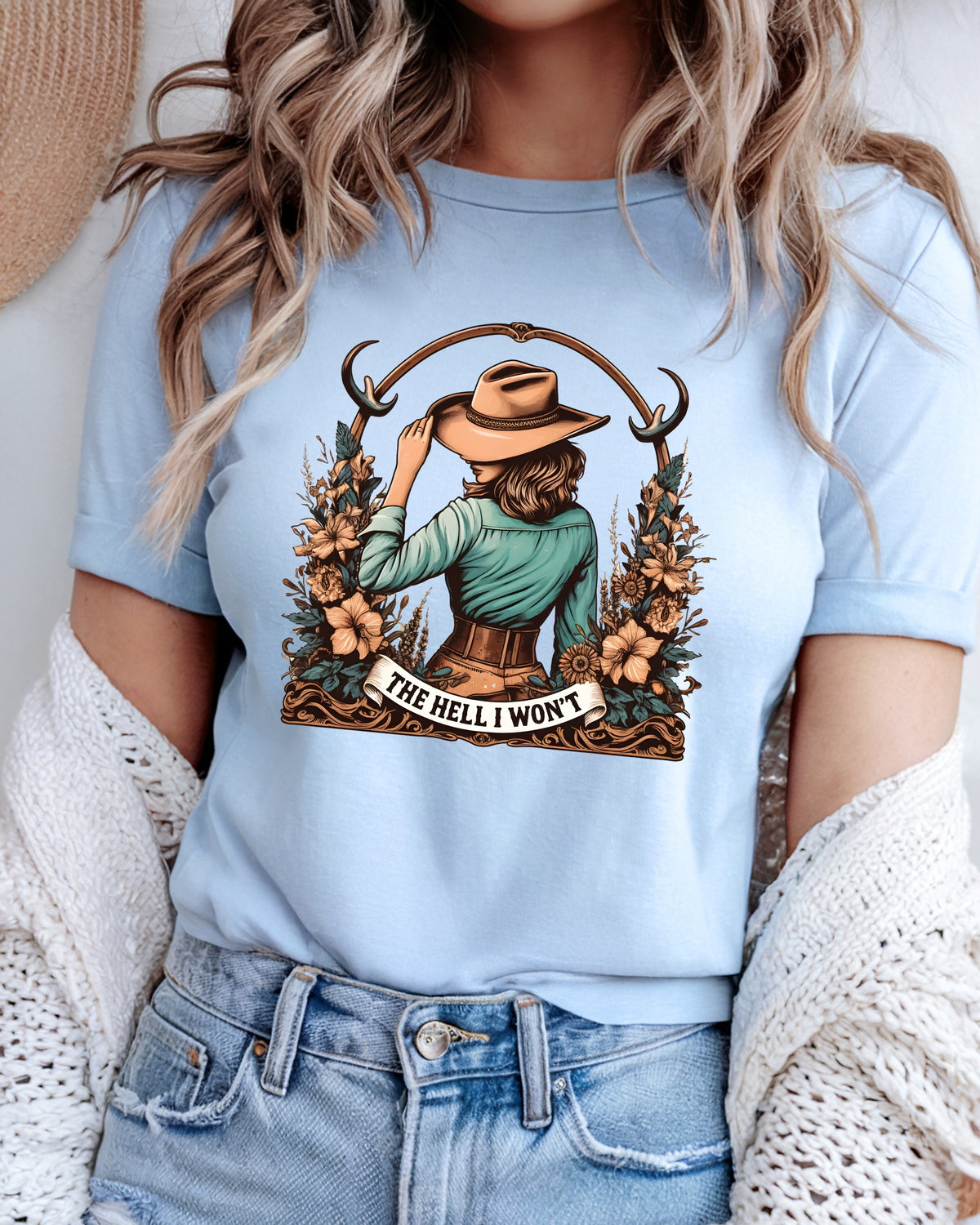 A woman wearing a light blue graphic t-shirt with a western-themed illustration of a woman in a hat and the text 'THE HELL I WON'T'.