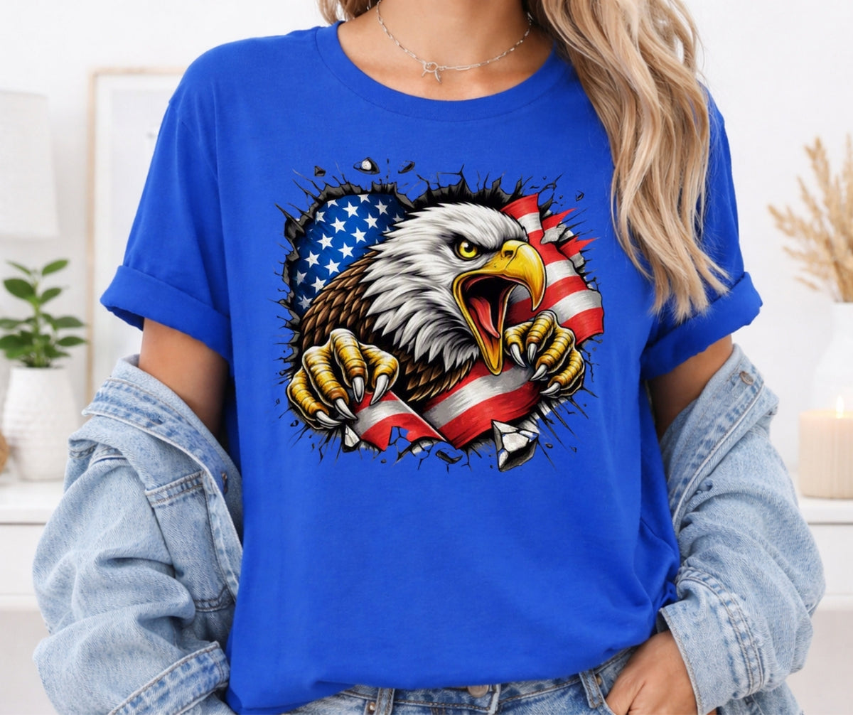 A woman wearing a bright blue short-sleeve t-shirt with a graphic of a bald eagle tearing through an American flag. The t-shirt is styled with a light-wash denim jacket draped off the shoulders, set against a softly blurred indoor background.