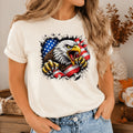 A woman wearing a cream-colored t-shirt with a graphic of a bald eagle tearing through an American flag. The background shows a cozy indoor setting with a vase and autumn decorations.
