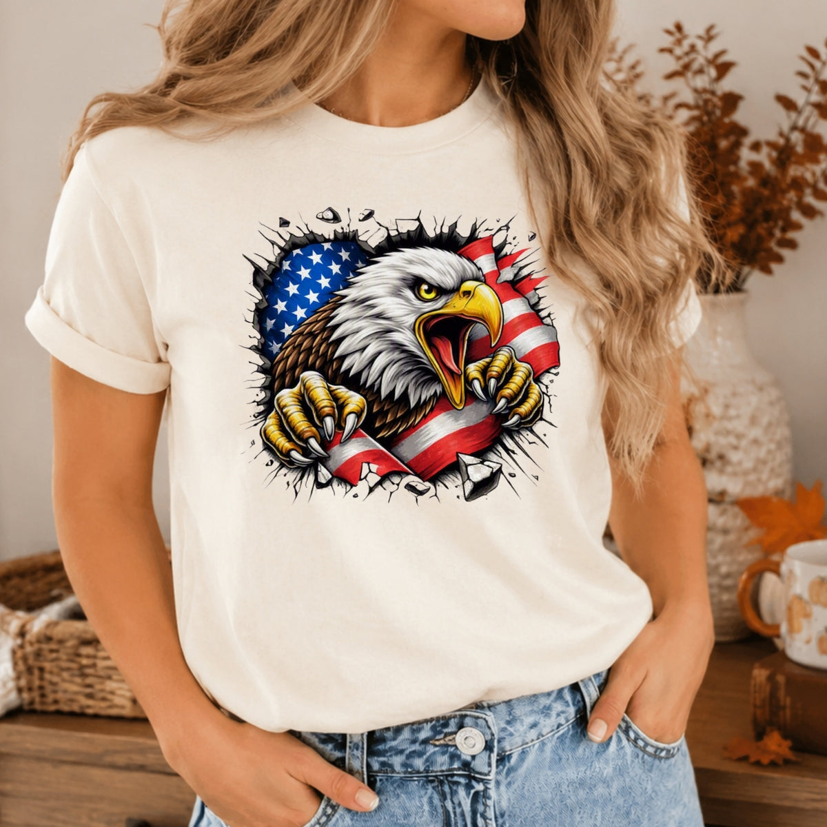 A woman wearing a cream-colored t-shirt with a graphic of a bald eagle tearing through an American flag. The background shows a cozy indoor setting with a vase and autumn decorations.