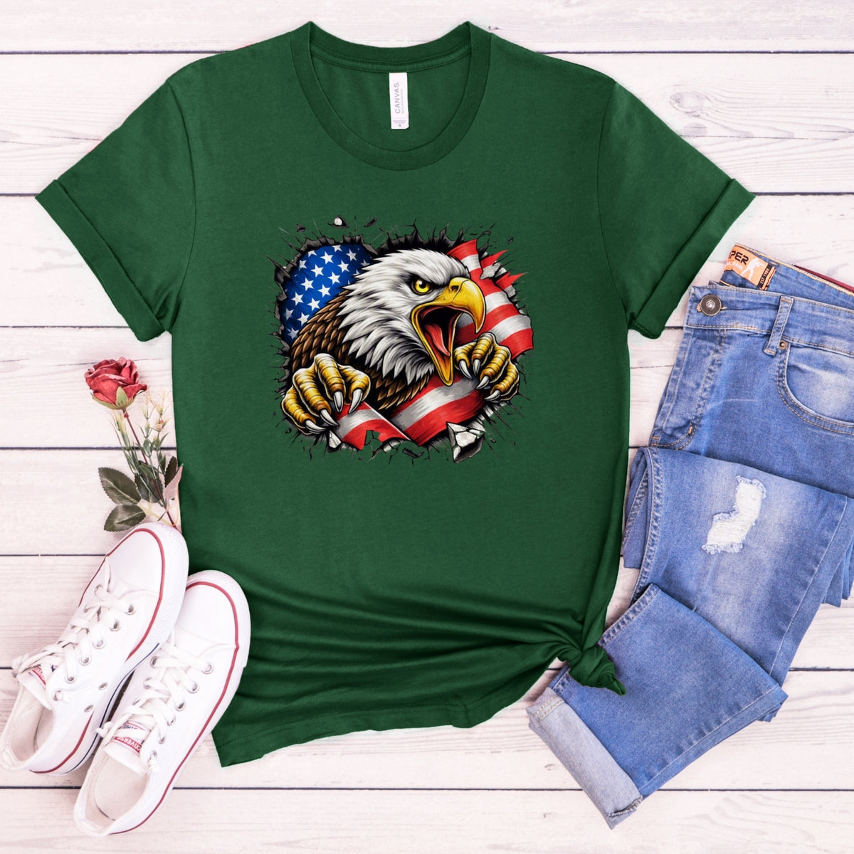 A green short-sleeve t-shirt with a graphic of a bald eagle tearing through an American flag, styled in a flat lay with blue jeans, white sneakers, and a single red rose on a white wood plank background.