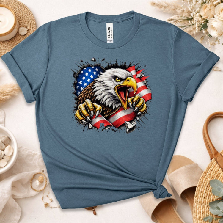A slate blue crewneck t-shirt featuring a graphic of a bald eagle tearing through an American flag. The shirt is styled in a flat-lay arrangement with neutral props including a straw hat, a small bowl of shells, and beige sandals on a light beige background.