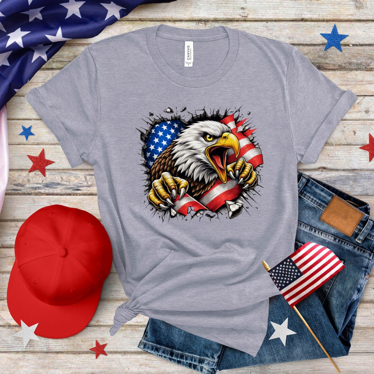 A heather gray graphic t-shirt featuring a bald eagle tearing through an American flag design, styled in a flat lay with blue jeans, a red baseball cap, a small American flag, and patriotic star decorations on a rustic wood background.
