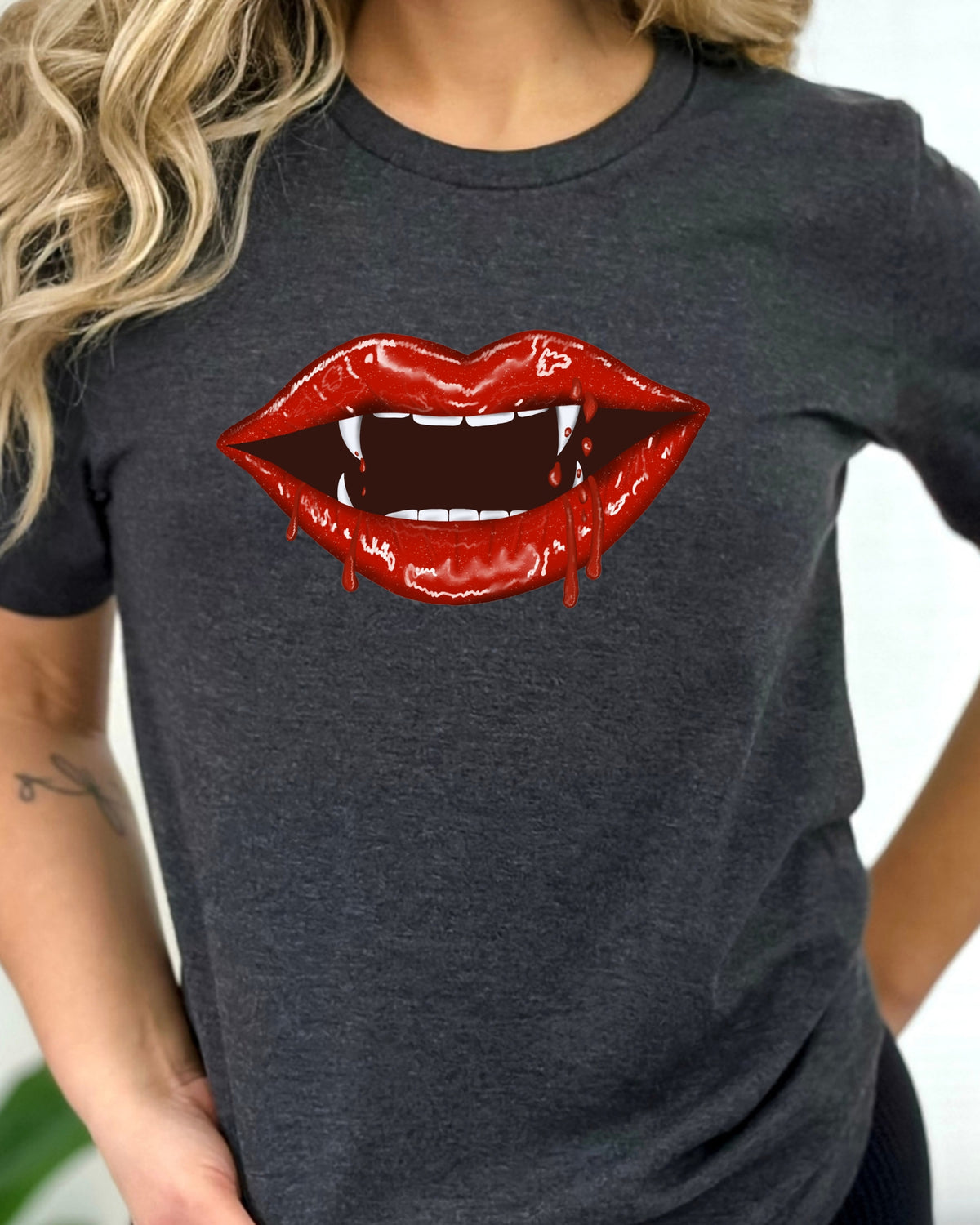 A woman wearing a dark heather gray t-shirt with a graphic of glossy red vampire lips showing fangs.