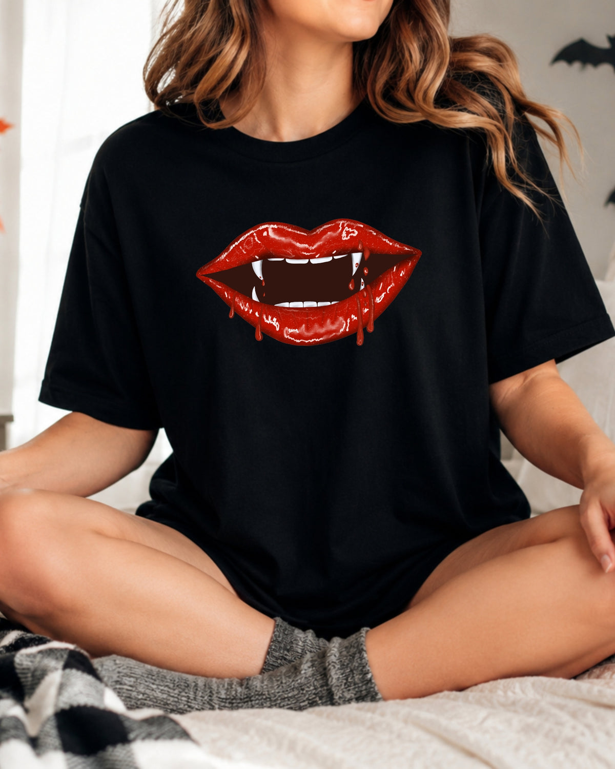 A woman sitting cross-legged on a bed wearing a black t-shirt with a graphic of red vampire lips with white fangs.