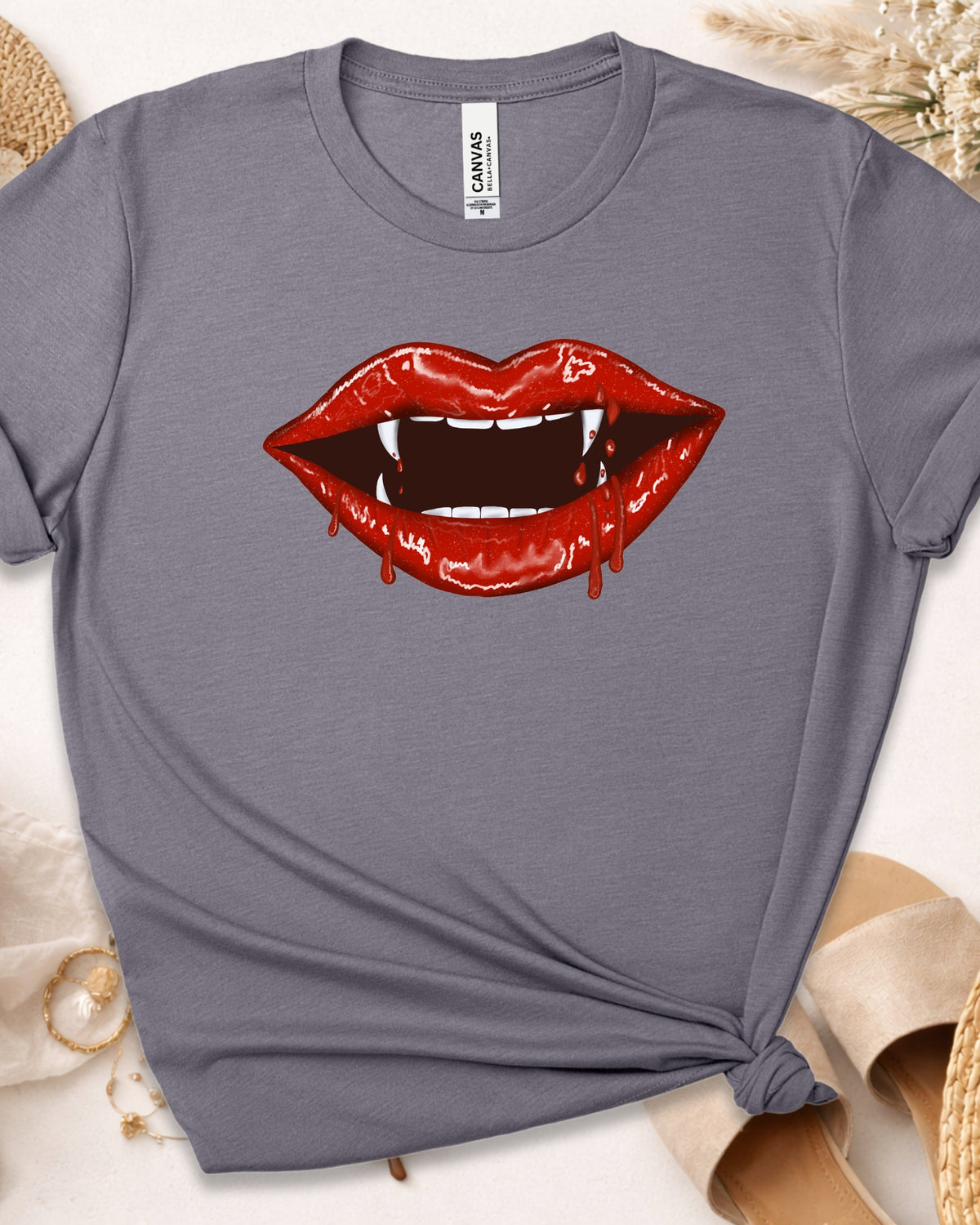 A heather gray short-sleeve t-shirt with a graphic of vampire fangs and dripping red lips on the front. The shirt is styled flat on a light beige background with dried flowers and sandals.