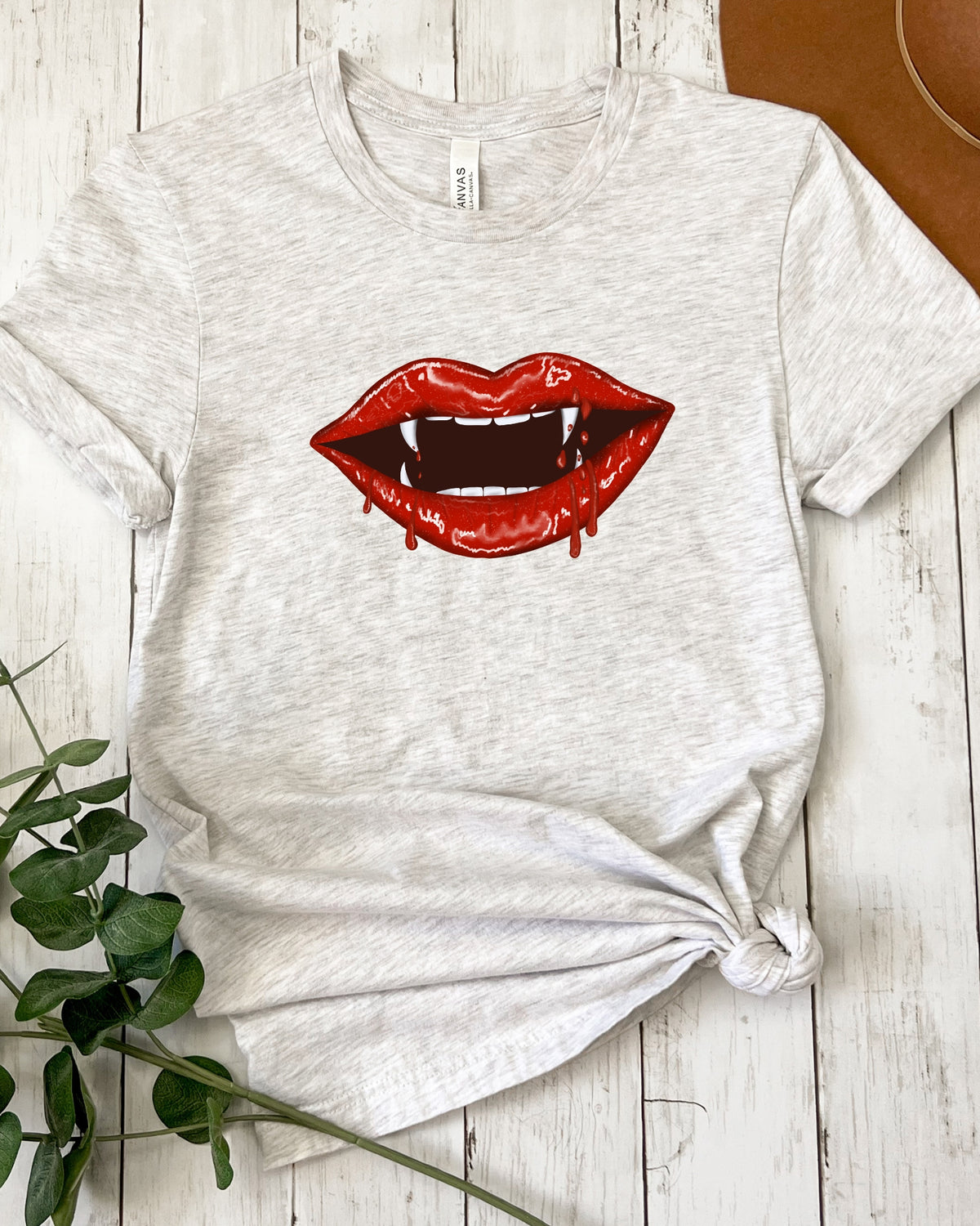 A light heather gray t-shirt with a graphic of dripping red vampire lips on the front, styled in a flat-lay on a white wood plank background with a green plant and a brown hat visible.