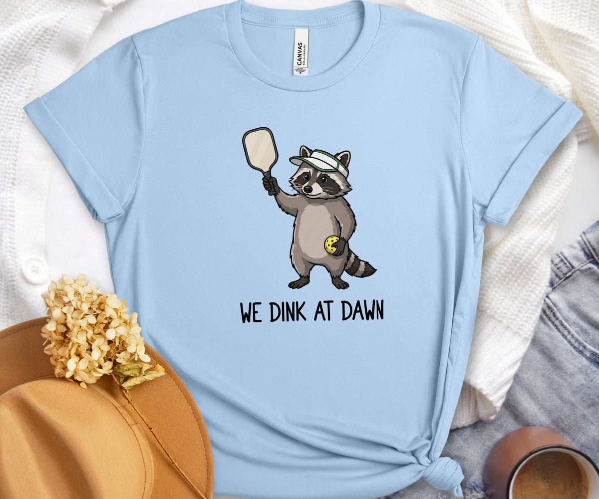 A light blue short-sleeve t-shirt with a cartoon raccoon holding a paddle and the text 'WE DINK AT DAWN' printed on the front. The shirt is styled in a flat-lay arrangement with a tan hat, dried flowers, and denim on a white textured background.