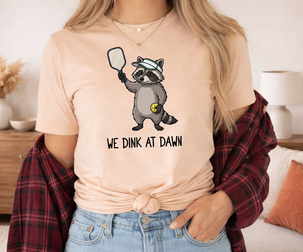 A woman wearing a light peach t-shirt with a cartoon raccoon graphic holding a mirror and the text 'WE DINK AT DAWN'. She has a red plaid flannel shirt tied around her shoulders and is wearing light blue jeans in a cozy indoor setting.