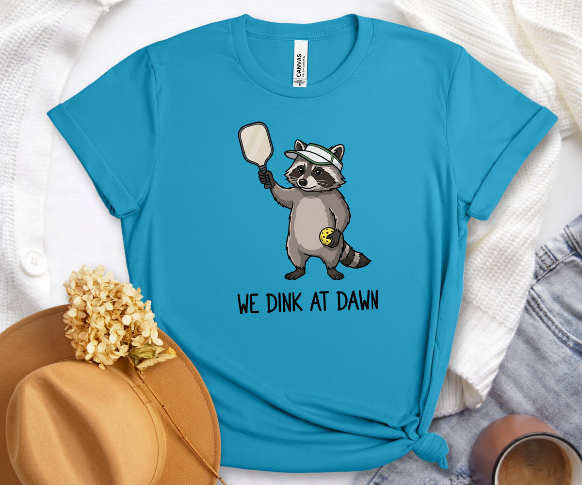 A bright turquoise blue t-shirt with a cartoon raccoon holding a paddle and the text 'WE DINK AT DAWN' printed on the front. The shirt is styled in a flat-lay arrangement with a tan hat, white knit fabric, and denim.
