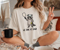 A woman sitting cross-legged on a bed wearing an oversized cream graphic t-shirt with a cartoon raccoon holding a mirror and the text 'WE DINK AT DAWN'. She is holding a black mug and making a peace sign.