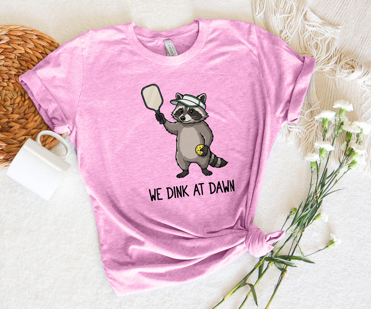 A pink short-sleeve graphic t-shirt laid flat on a white textured fabric background, styled with a woven mat, a white mug, and white flowers. The shirt features a cartoon raccoon holding a mirror with the text 'WE DINK AT DAWN' underneath.