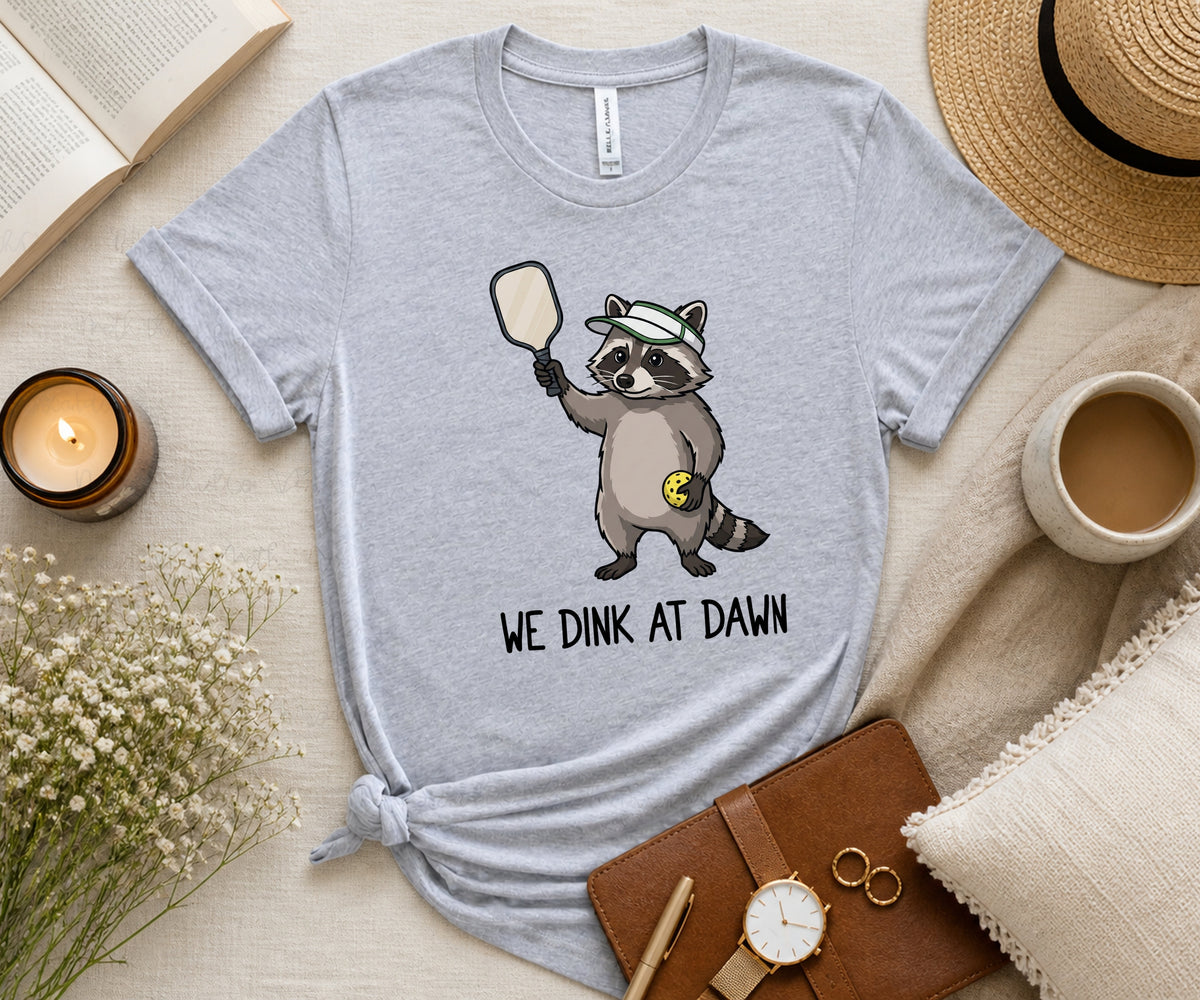 A light heather gray graphic t-shirt featuring a cartoon raccoon holding a mirror and the text 'WE DINK AT DAWN'. The t-shirt is styled in a cozy flat-lay arrangement with a straw hat, a cup of coffee, a candle, a notebook, and a watch on a beige fabric background.