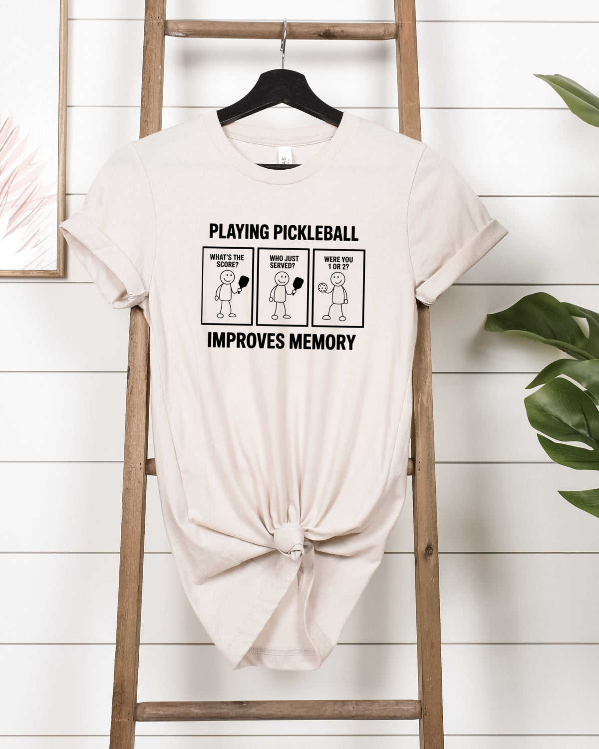A light beige graphic t-shirt with a black printed design that reads 'PLAYING PICKLEBALL IMPROVES MEMORY' with three cartoon panels, hanging on a wooden hanger on a rustic wooden ladder against a white shiplap wall.