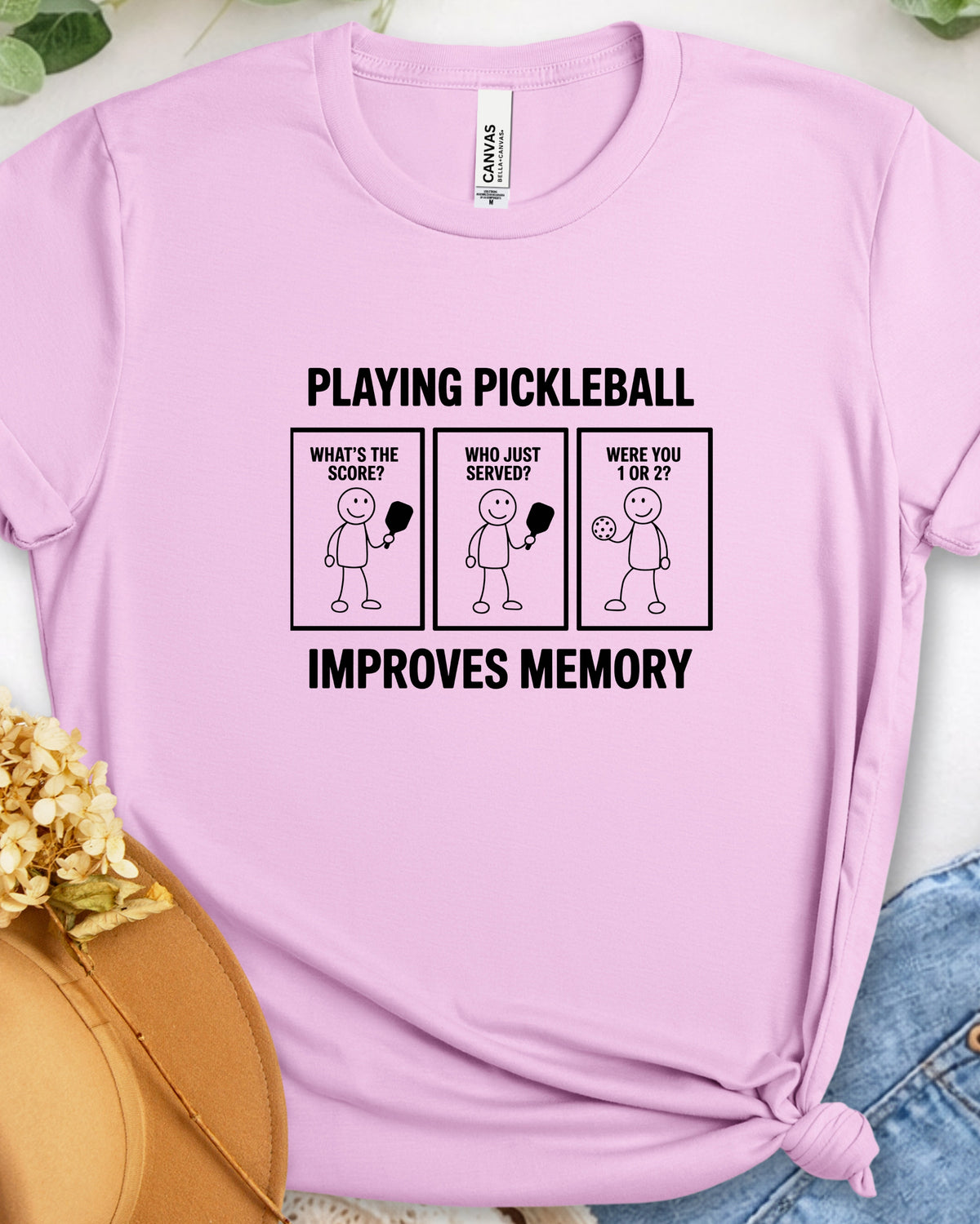 A light pink short-sleeve t-shirt with a black graphic print on the front that reads 'PLAYING PICKLEBALL IMPROVES MEMORY' with three cartoon panels. The shirt is styled flat on a white surface with a straw hat, dried flowers, and blue jeans partially visible.