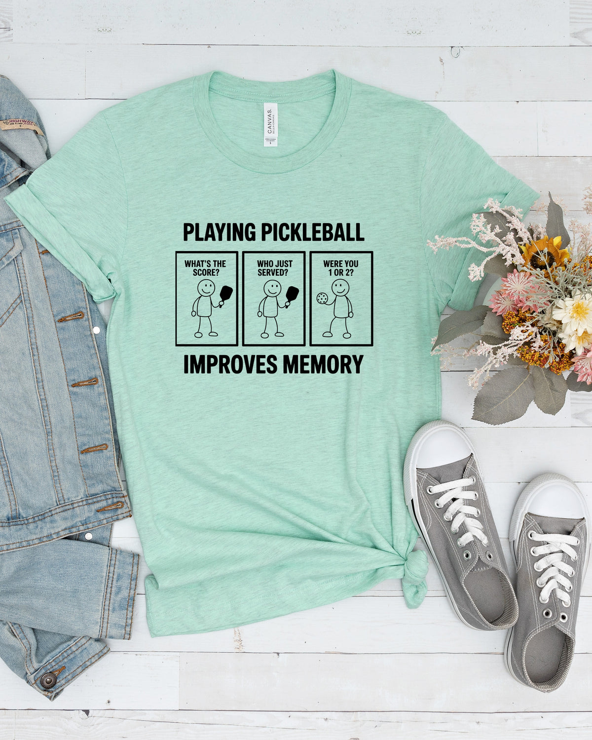 A mint green graphic t-shirt featuring a pickleball-themed memory mnemonic, styled in a flat lay with a denim jacket, gray sneakers, and a small bouquet of flowers on a white wood background.