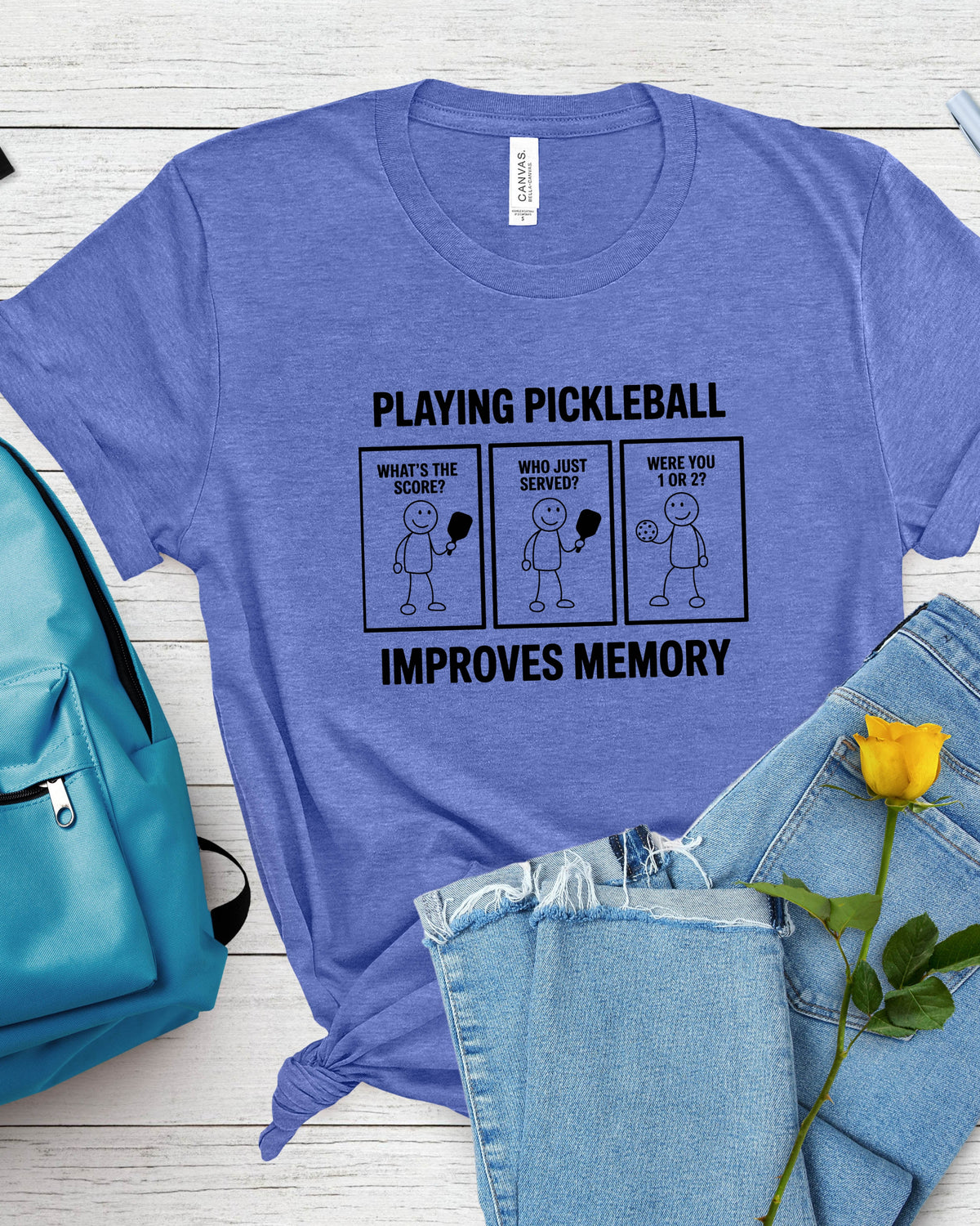 A heathered periwinkle blue t-shirt with a black graphic print about pickleball and memory, styled in a flat lay with light-wash denim shorts, a turquoise backpack, and a yellow rose on a white wood background.