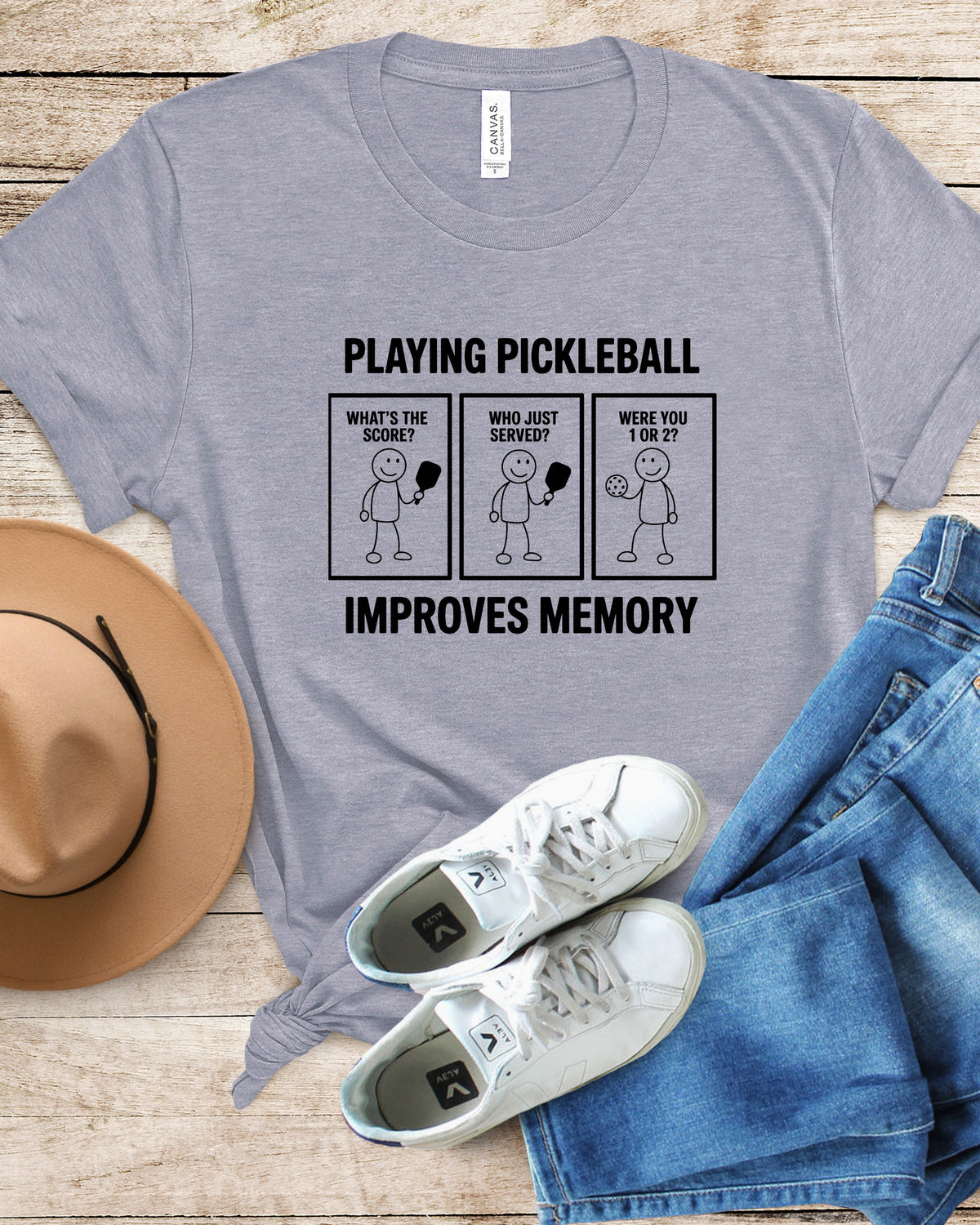 A heather gray graphic t-shirt laid flat on a light wood background, styled with a tan hat, white sneakers, and blue jeans. The t-shirt features a black printed graphic with three stick-figure panels and the text 'PLAYING PICKLEBALL IMPROVES MEMORY'.