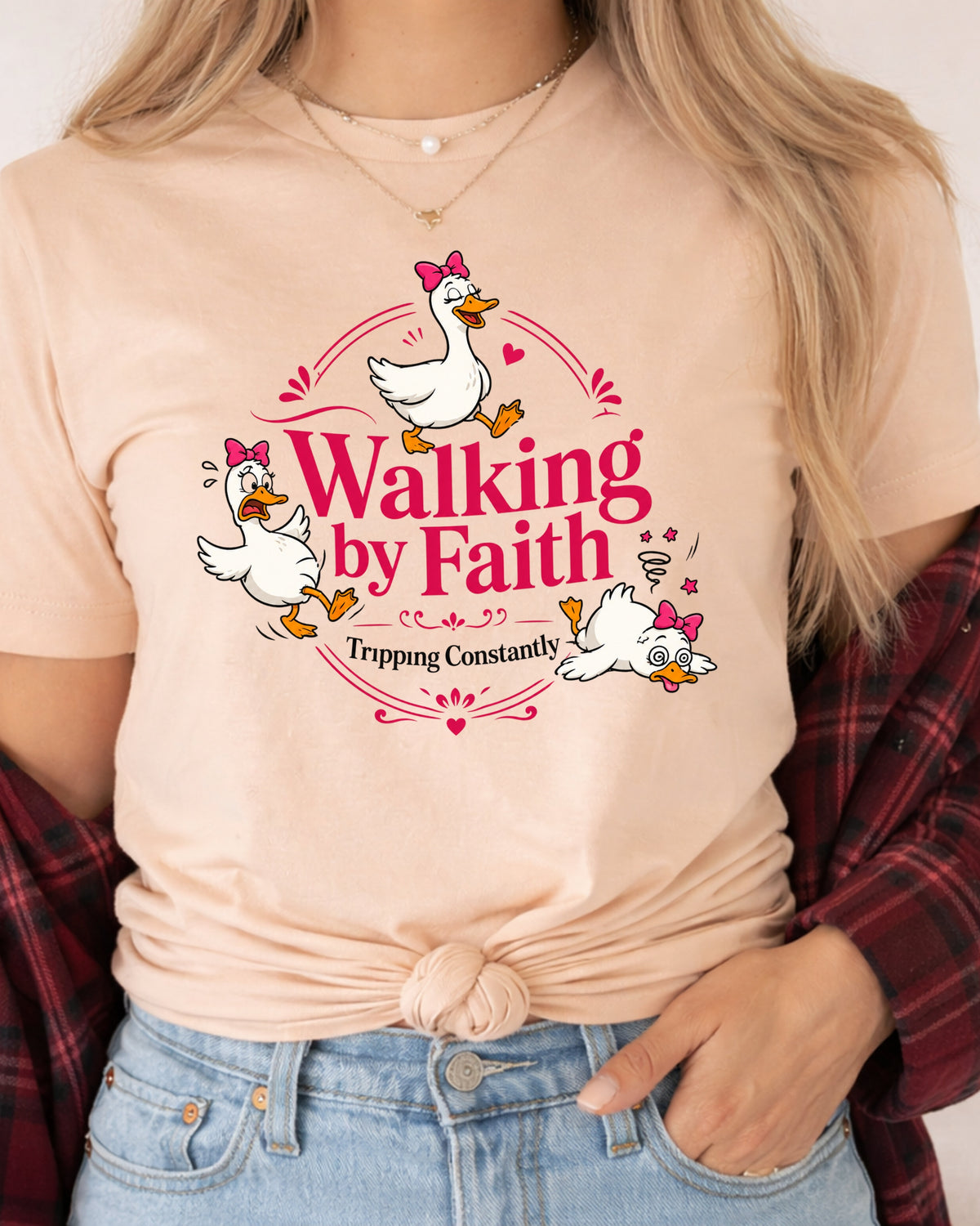 A woman wearing a light peach t-shirt with a cartoon graphic of three white ducks with pink bows and the text 'Walking by Faith Tripping Constantly'. She has a red plaid shirt tied off her shoulders and is wearing blue jeans.