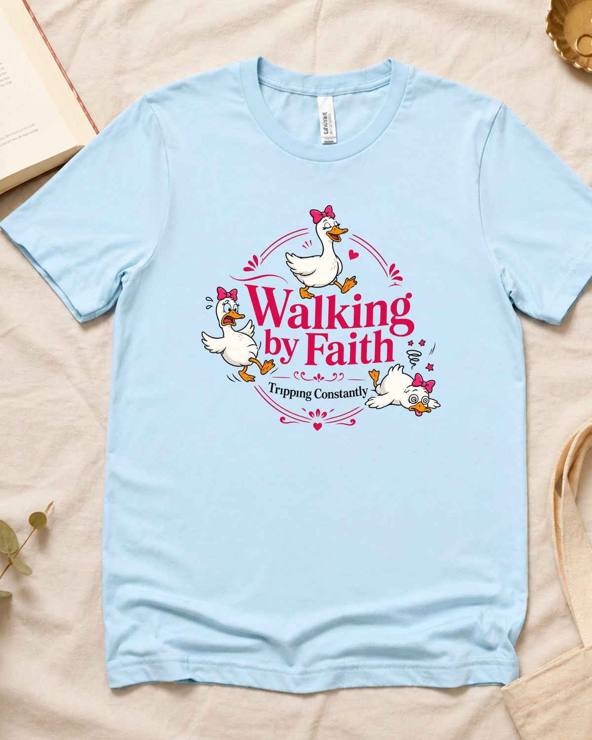 A light blue short-sleeve t-shirt laid flat on a beige fabric background, featuring a graphic print of three cartoon white geese with pink bows and the text 'Walking by Faith' and 'Trusting Constantly'.