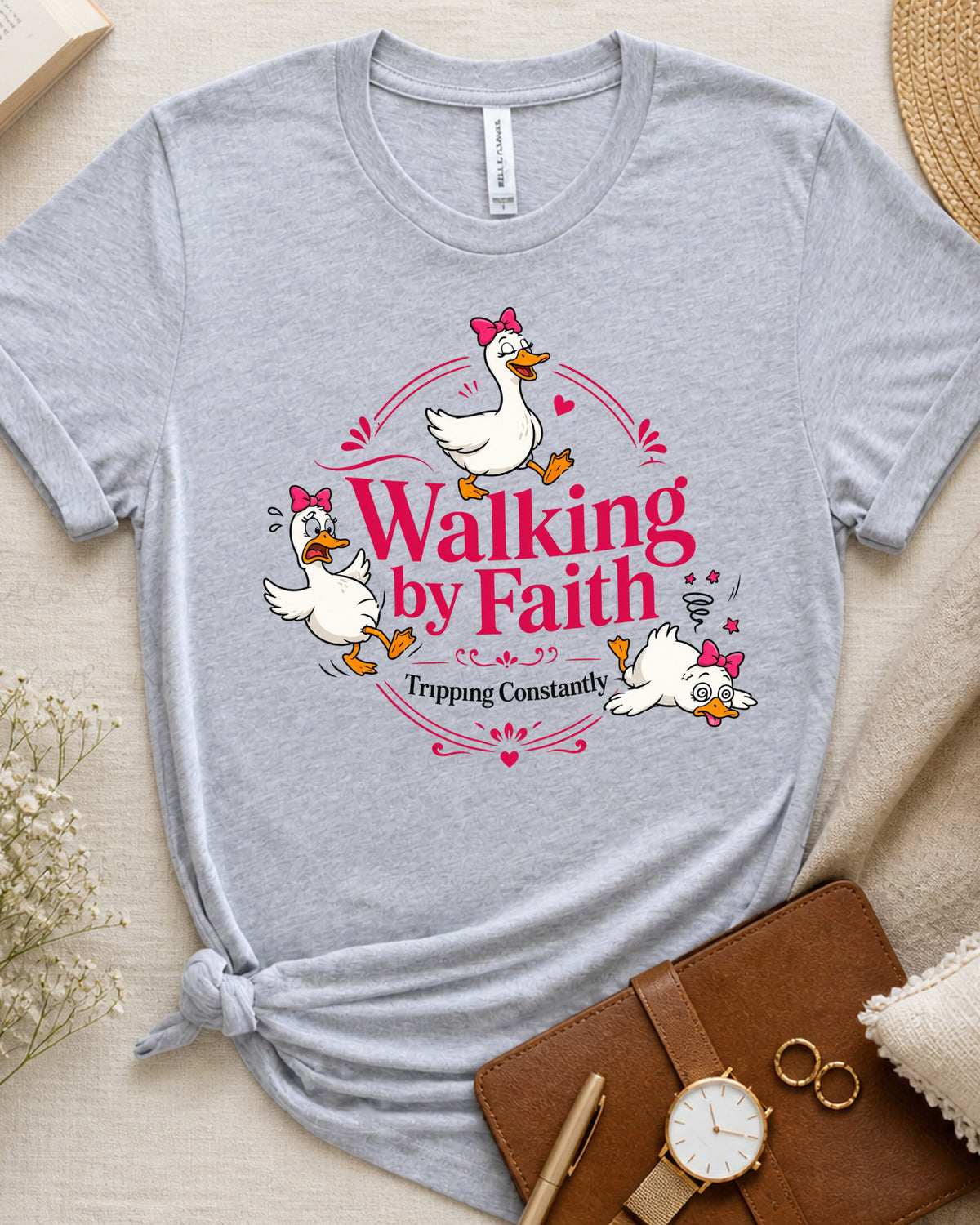 A light heather gray t-shirt with a graphic of three white ducks wearing pink bows, walking in a circle with the text 'Walking by Faith Trippong Constantly' in pink and white. The t-shirt is styled in a flat lay with a brown leather notebook, a watch, and dried flowers on a beige fabric background.