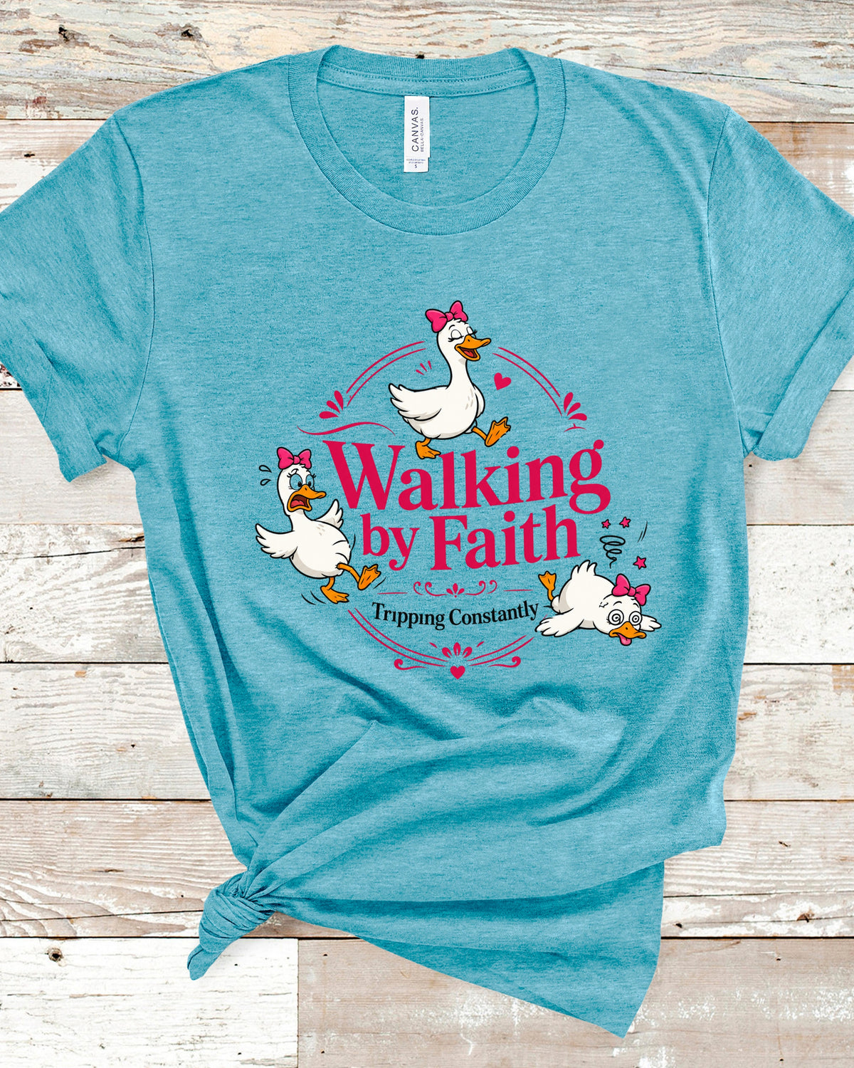 A heathered teal t-shirt with a graphic print of three cartoon ducks and the text 'Walking by Faith Tripping Constantly', displayed flat on a light wood plank background.
