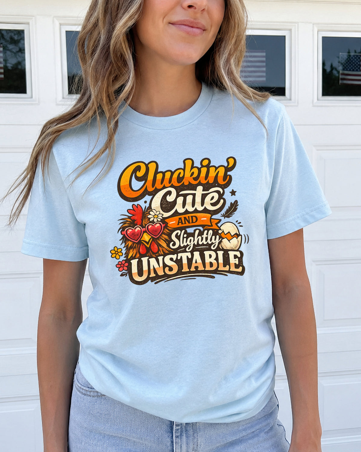 A woman wearing a light blue graphic t-shirt with the text 'Cluckin' Cute and Slightly UNSTABLE' in a rustic orange, yellow, and brown font, paired with light-wash jeans, standing in front of a white garage door.